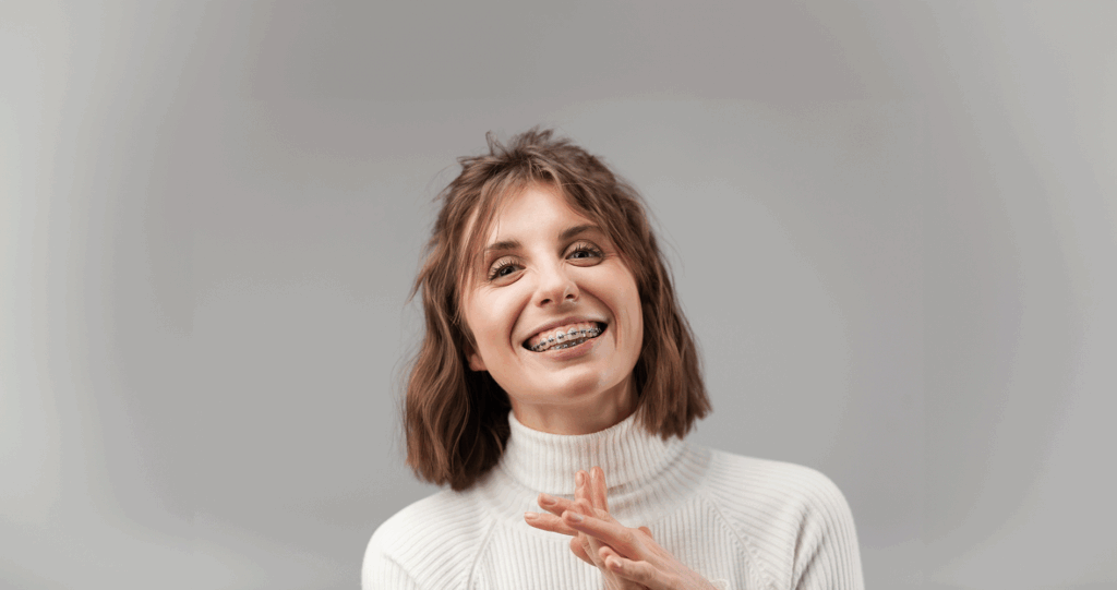 Adult Woman clasping her hands and smiling in front of a gray backdrop while sporting her braces
