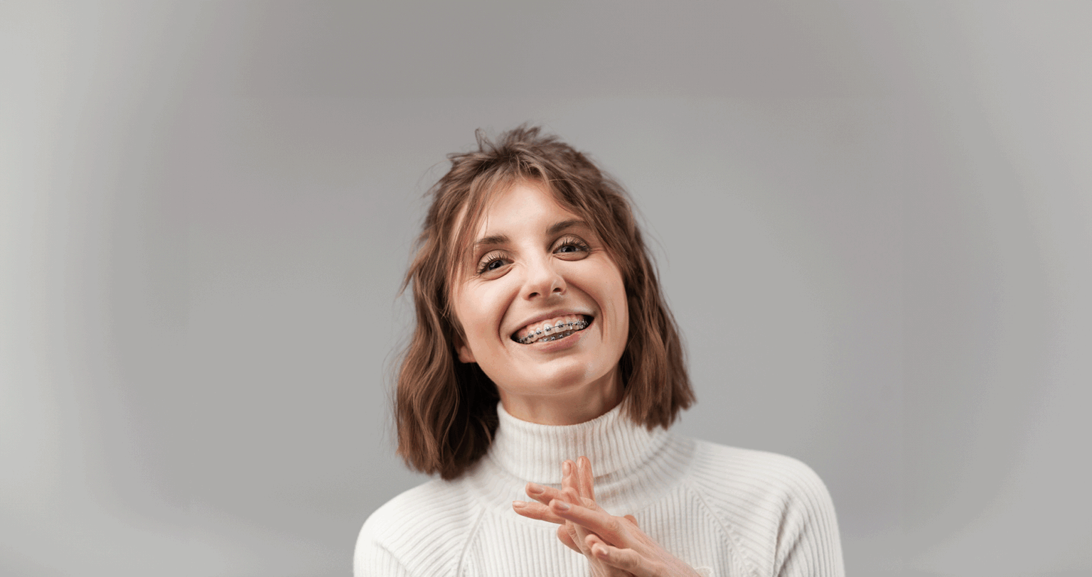 Adult Woman clasping her hands and smiling in front of a gray backdrop while sporting her braces
